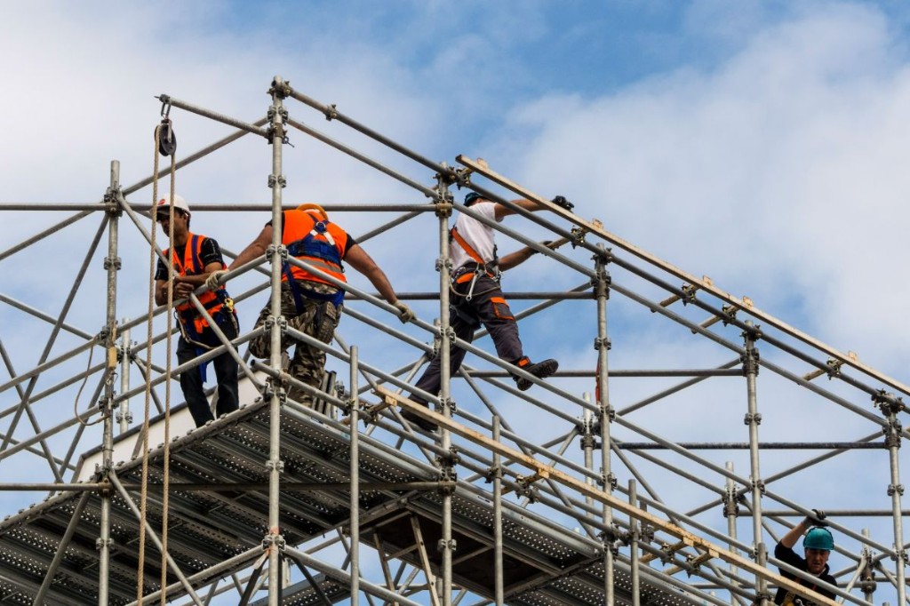 Workers in harnesses assembling and working on industrial scaffolding against a blue sky